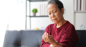 Older woman sits on a sofa, examining her wrist with a pained expression in a living room.
